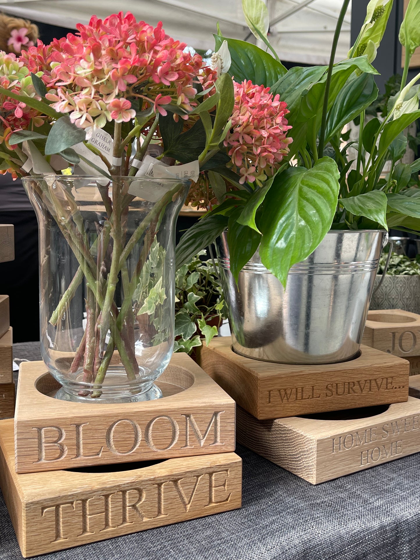 Decorative wooden oak plant pot stands with floral arrangements.  Wooden plant pot stands individually engraved with "BLOOM" "THRIVE" "I WILL SURVIVE" HOME SWEET HOME" 