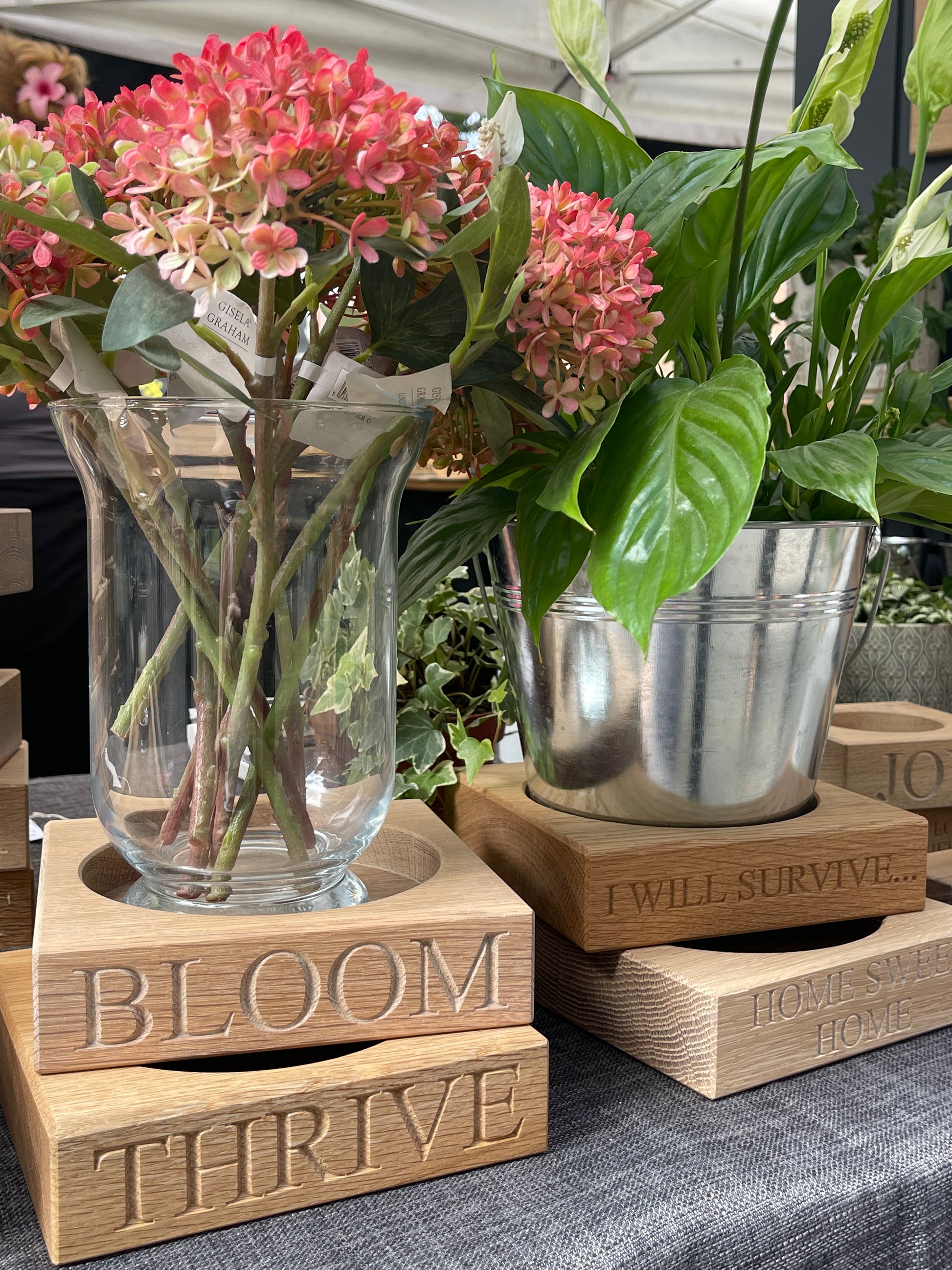 Decorative wooden oak plant pot stands with floral arrangements.  Wooden plant pot stands individually engraved with "BLOOM" "THRIVE" "I WILL SURVIVE" HOME SWEET HOME" 