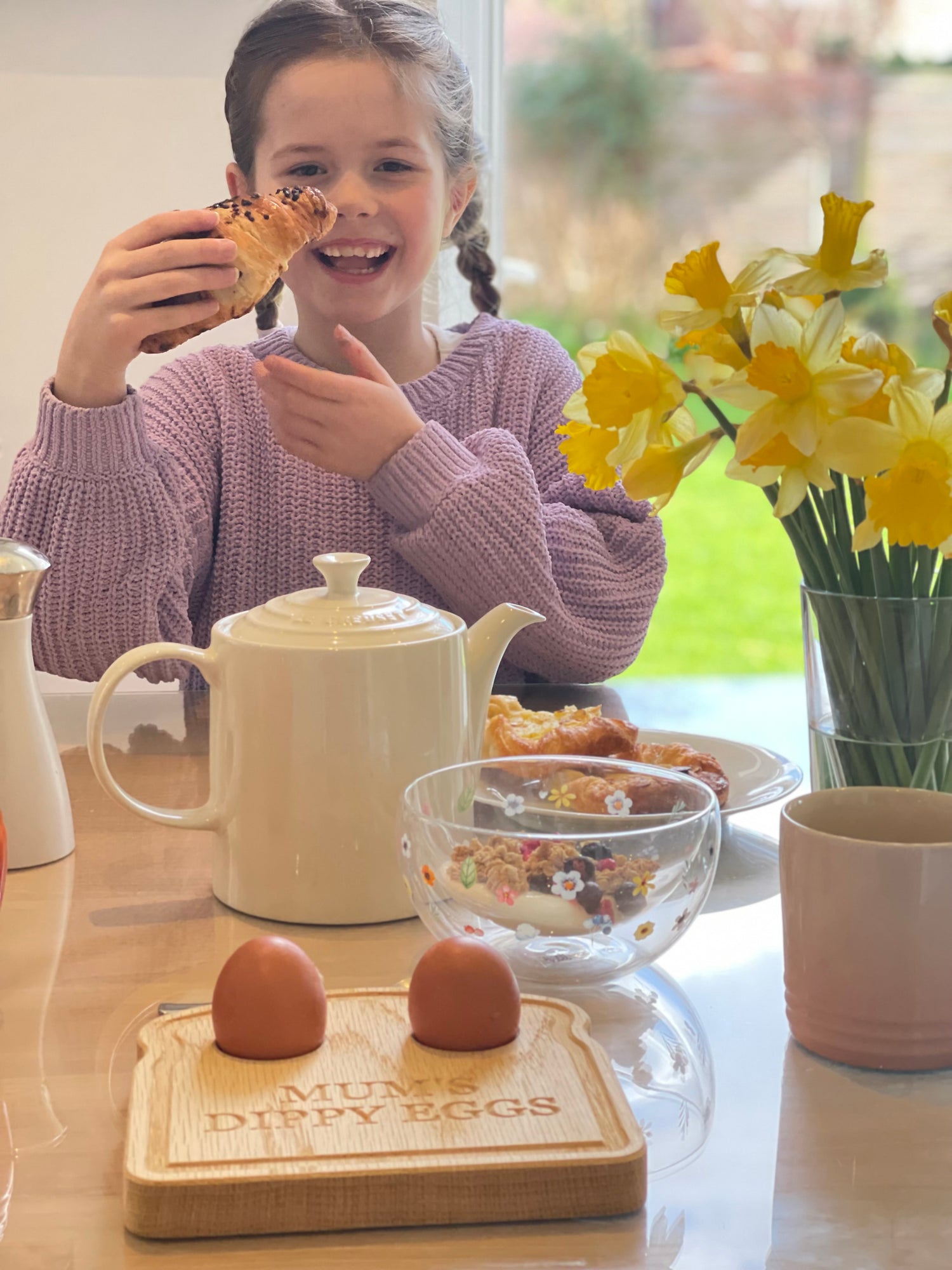 Child eating a breakfast at a table with a teapot, toast shaped oak egg plate, and flowers.